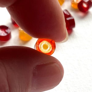 May include: A close-up of a single red and orange translucent candy. The candy is round and has a small hole in the center. The candy is being held between two fingers.