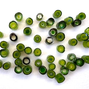 May include: A scattering of small, cylindrical glass beads in shades of green and clear. The beads have a hollow centre and are spread across a white surface. The beads vary in size and shape, with some appearing more opaque than others.