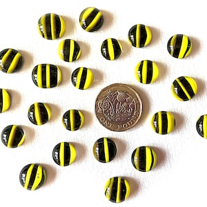 May include: A scattering of yellow and black striped glass marbles on a white background. A British one-pound coin is included for scale. The marbles are round and have a glossy finish, resembling miniature bumblebees.
