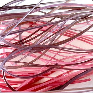 May include: A bundle of thin, flexible glass rods in shades of pink, purple, and brown. The rods are arranged in a random, tangled pattern.