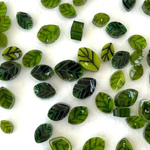 Peut inclure: Collection de petites perles de verre en forme de feuilles, dans des tons de vert. Chaque perle présente des détails de nervures vert foncé, créant un aspect de feuille réaliste. Les perles sont dispersées sur une surface blanche.