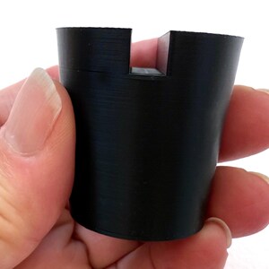 May include: A black, cylindrical object with a rectangular notch cut out of the top. The object is held in a hand against a white background. The object appears to be made of a solid, matte material.