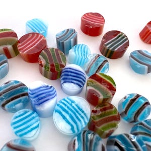 May include: A selection of colourful, striped hard candies in shades of red, green, blue, and brown. The candies are cylindrical and oval, with a glossy finish. The candies are scattered on a white surface.
