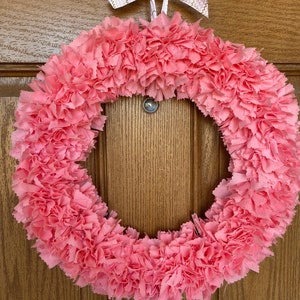 May include: A pink fabric wreath with a circular design. The wreath is made of layered, ruffled fabric, creating a textured appearance. A white ribbon with gold zigzag patterns is tied at the top for hanging. The wreath is displayed against a wooden door.