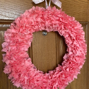 May include: A handmade pink fabric wreath with a circular design. The wreath is made of layered, ruffled fabric, creating a textured appearance. It is suspended by a white hook and a pink ribbon with gold zigzag patterns.