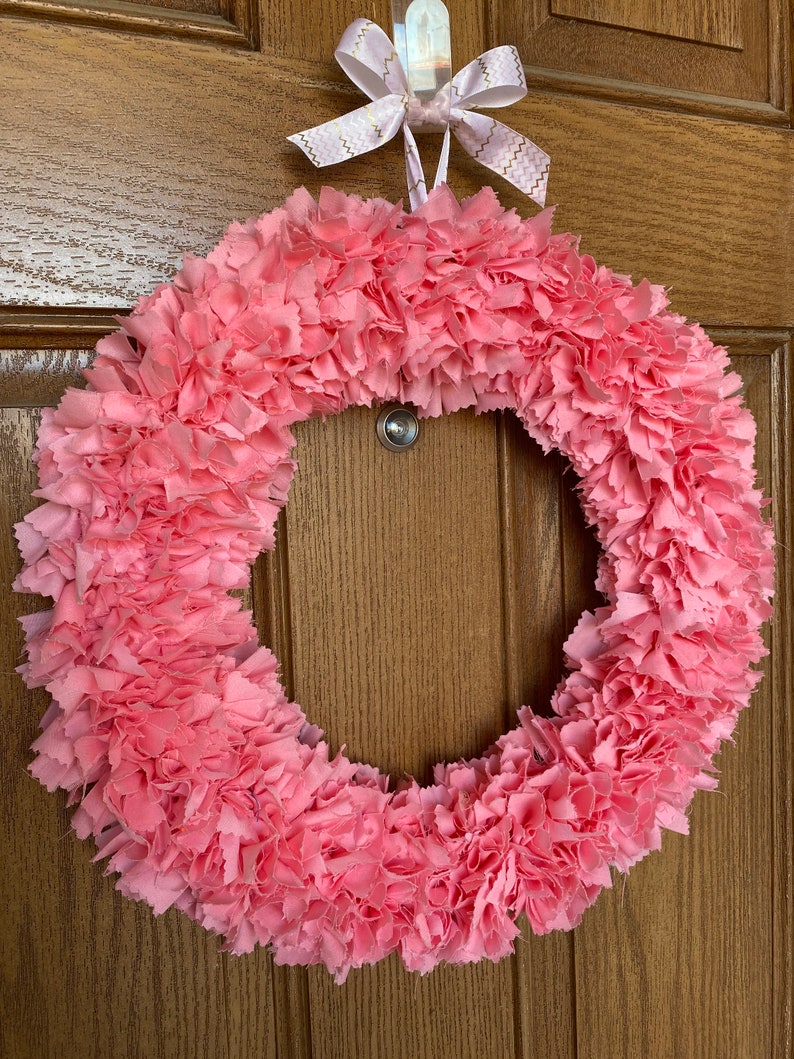 May include: A handmade pink fabric wreath with a circular design. The wreath is made of layered, ruffled fabric, creating a textured appearance. It is suspended by a white hook and a pink ribbon with gold zigzag patterns.