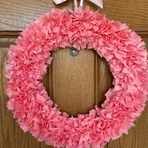 May include: A pink fabric wreath with a circular design. The wreath is made of layered, ruffled fabric, creating a textured appearance. A white ribbon with gold zigzag patterns is tied at the top for hanging. The wreath is displayed against a wooden door.