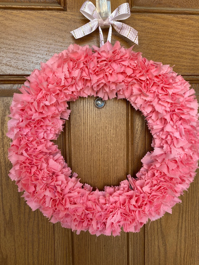 May include: A pink fabric wreath with a circular design. The wreath is made of layered, ruffled fabric, creating a textured appearance. A white ribbon with gold zigzag patterns is tied at the top for hanging. The wreath is displayed against a wooden door.