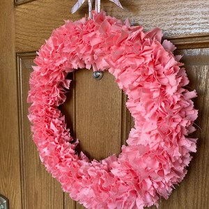 May include: A handmade pink fabric wreath with a circular design. The wreath is made of layered, ruffled fabric and is suspended by a white ribbon with gold accents. The wreath is hanging on a wooden door.