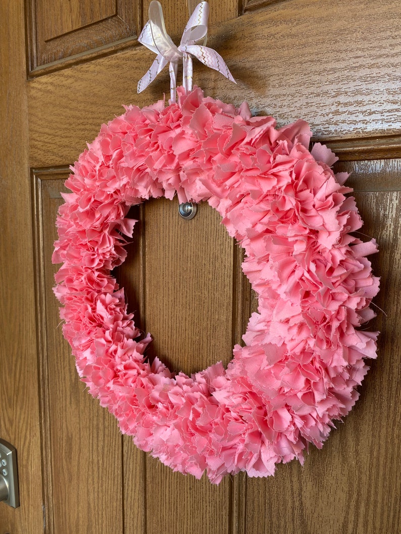 May include: A handmade pink fabric wreath with a circular design. The wreath is made of layered, ruffled fabric and is suspended by a white ribbon with gold accents. The wreath is hanging on a wooden door.