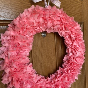 May include: A handmade pink fabric wreath with a circular design. The wreath is made of layered, ruffled fabric, creating a textured appearance. A white ribbon with gold zigzag patterns is tied at the top for hanging. The wreath is displayed on a wooden door.