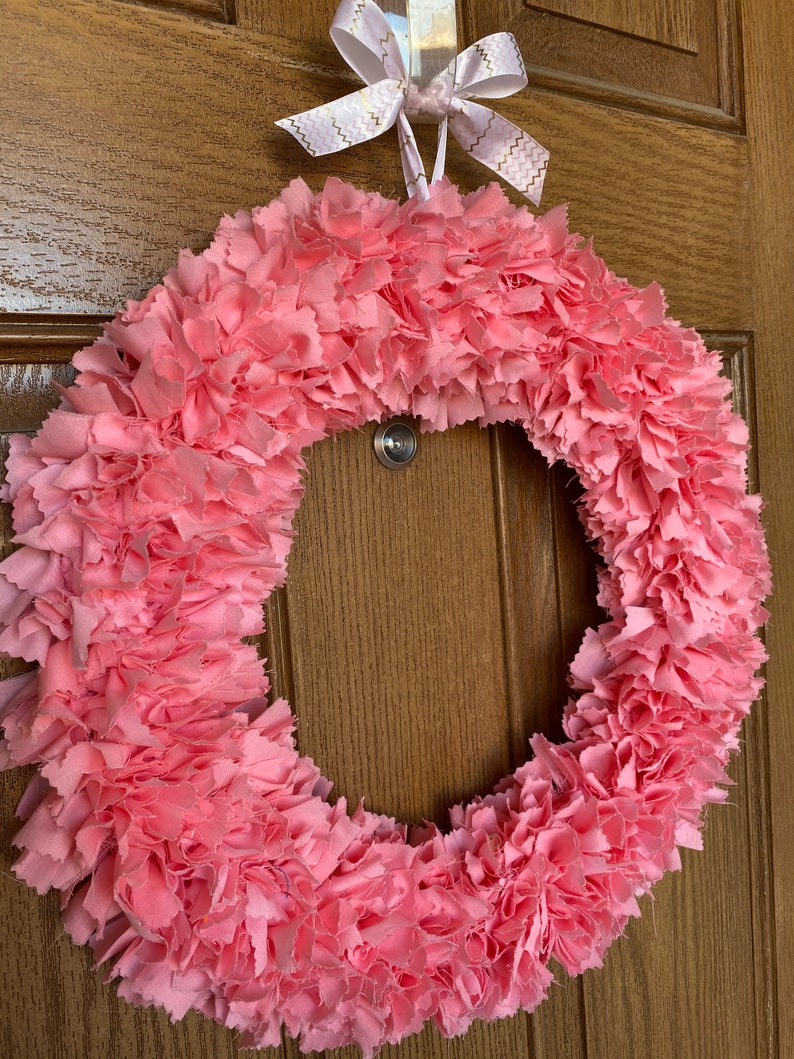 May include: A handmade pink fabric wreath with a circular design. The wreath is made of layered, ruffled fabric, creating a textured appearance. A white ribbon with gold zigzag patterns is tied at the top for hanging. The wreath is displayed on a wooden door.