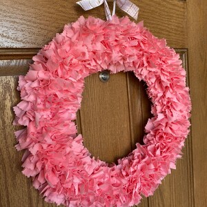 May include: A handmade pink fabric wreath with a circular design. The wreath is made of layered, ruffled fabric, creating a textured appearance. A white and gold ribbon bow is attached at the top for hanging. The wreath is displayed on a wooden door.