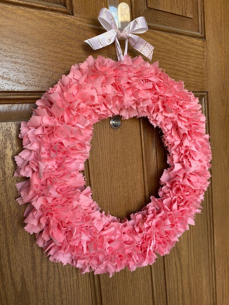 May include: A handmade pink fabric wreath with a circular design. The wreath is made of layered, ruffled fabric, creating a textured appearance. A white and gold ribbon bow is attached at the top for hanging. The wreath is displayed on a wooden door.