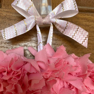 May include: A handmade pink fabric wreath with a white and gold chevron patterned ribbon bow. The wreath is made of layered, ruffled fabric, creating a textured, floral appearance. The bow is tied at the top for hanging.