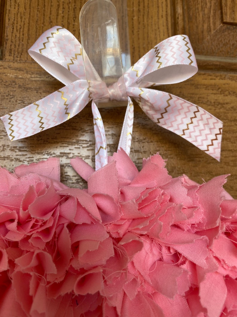 May include: A handmade pink fabric wreath with a white and gold chevron patterned ribbon bow. The wreath is made of layered, ruffled fabric, creating a textured, floral appearance. The bow is tied at the top for hanging.