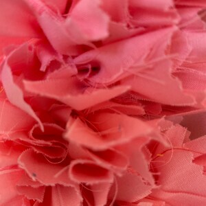 May include: Close-up of a soft, pink fabric with a layered, ruffled texture. The fabric appears to be cut into strips, creating a textured, floral-like design. The color is a light, muted pink.