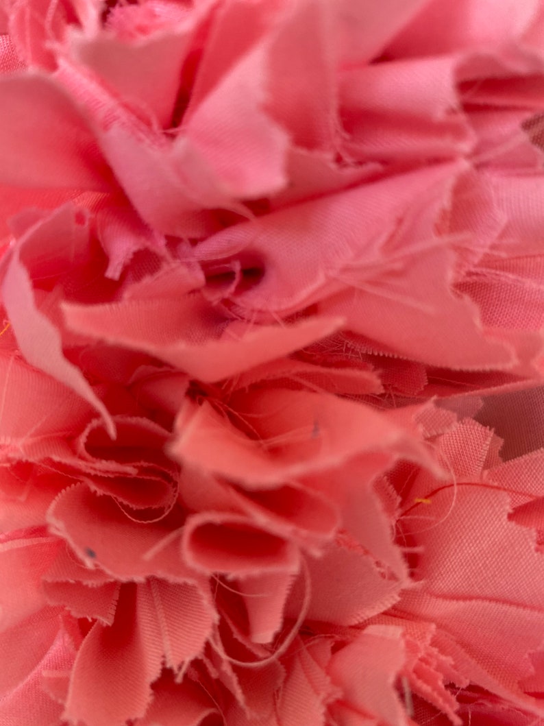 May include: Close-up of a soft, pink fabric with a layered, ruffled texture. The fabric appears to be cut into strips, creating a textured, floral-like design. The color is a light, muted pink.