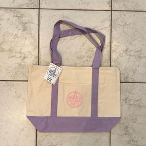 May include: Cream-colored canvas tote bag with lavender accents. Features a front pocket and lavender handles. The bag has a lavender bottom and a pink Trader Joe's logo. A Trader Joe's tag is attached to the handle.