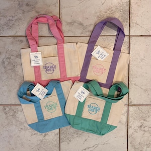 May include: Four mini pastel canvas tote bags. The bags are in pink, purple, blue, and teal. Each bag has a cream-colored body with a matching colored handle and bottom. The Trader Joe's logo is printed on the front of each bag.