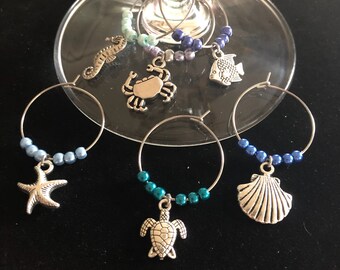 May include: Set of six sea-themed wine glass charms. Each charm features a different sea creature, including a starfish, seahorse, crab, turtle, seashell, and fish. The charms are made of silver metal and are strung on a wire ring with blue, green, and purple beads.