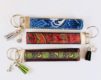 May include: Three keychains with patterned fabric straps in blue, red, and green. Each keychain has a silver-tone metal clasp and a tassel. The blue keychain has a silver tassel and a fish charm. The red keychain has a black tassel and a horseshoe charm. The green keychain has a green tassel and an anchor charm.