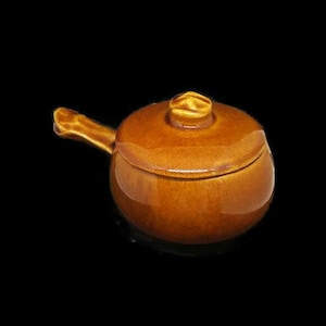 Puede incluir: Una pequeña olla redonda de cerámica marrón con tapa y asa. La olla tiene un acabado brillante y un diseño sencillo. El asa es curva y está unida al lateral. La tapa tiene un pequeño pomo en la parte superior.