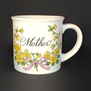 May include: White ceramic mug with a floral design featuring yellow flowers and green leaves. The mug has a pink ribbon and the word "Mother" written in black script.