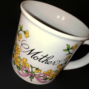 May include: White ceramic mug with a floral design featuring yellow flowers and green leaves. The mug has a pink ribbon and the word "Mother" written in black cursive.