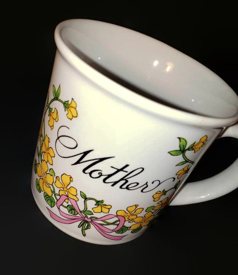 May include: White ceramic mug with a floral design featuring yellow flowers and green leaves. The mug has a pink ribbon and the word "Mother" written in black cursive.