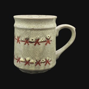May include: A beige ceramic mug with a speckled glaze and red stitched-like design. The mug has a rounded handle.