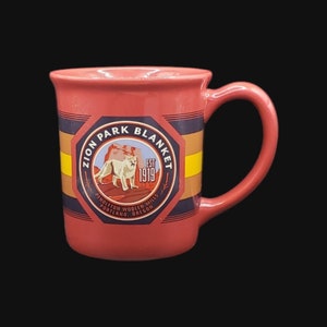 Könnte beinhalten: Rote Keramiktasse mit einem schwarz-gelben Streifenmuster. Die Tasse zeigt ein kreisförmiges Logo mit dem Text "Zion Park Blanket Est 1919 Pendleton Woolen Mills Portland Oregon" und einem stilisierten Bild eines Berglöwen.