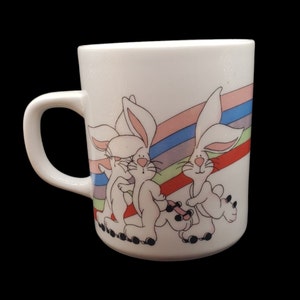 May include: White ceramic mug with a colorful rainbow design and three white rabbits on roller skates. The rabbits are wearing pink, blue, and green clothing.