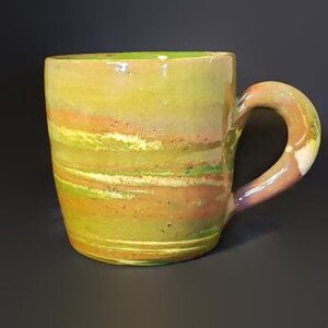 May include: A handmade ceramic mug with a green, yellow and orange glaze. The mug has a unique, swirled design and a rounded handle.