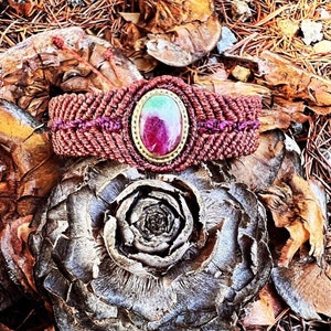 Peut inclure: Un bracelet tressé rouge et violet avec un sertissage doré orné d'une pierre précieuse verte et rose. Le bracelet est posé sur une surface brune texturée.