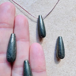 May include: Close-up of several teardrop-shaped, metallic gray beads. One bead is strung on a thin, brown cord. The beads have a textured surface and a slight sheen. The beads are held in a person's hand.