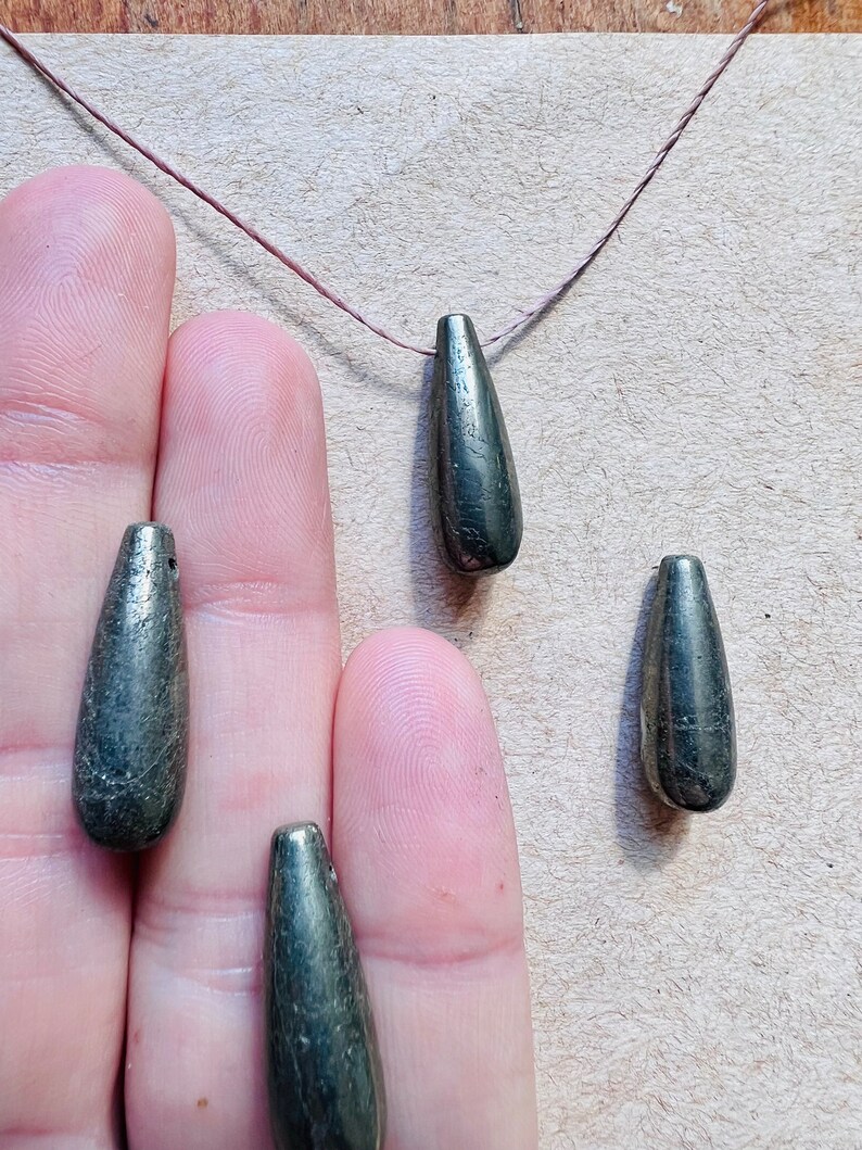 May include: Close-up of several teardrop-shaped, metallic gray beads. One bead is strung on a thin, brown cord. The beads have a textured surface and a slight sheen. The beads are held in a person's hand.