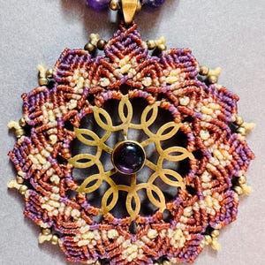 Puede incluir: Un collar con una piedra preciosa de amatista púrpura y un gran colgante de mandala intrincado hecho de cordón tejido y latón. El colgante presenta una piedra preciosa de amatista más pequeña en el centro.