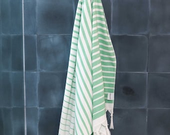 May include: A green and white striped towel with white fringe hanging on a hook in a bathroom. The towel has a small white tag with the word "Organic" printed on it.