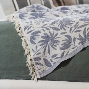 May include: A white throw blanket with a blue floral pattern and fringe edges. The blanket is draped over a green bedspread.