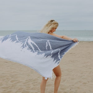 May include: A blue and white beach towel with a mountain design. The towel is being held up on a sandy beach. The towel has white fringe on the edges.