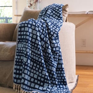 May include: A rectangular throw blanket featuring a repeating geometric pattern of dark navy blue and light blue squares. The textile has a textured surface and is finished with short, cream-colored fringe along the bottom edge.