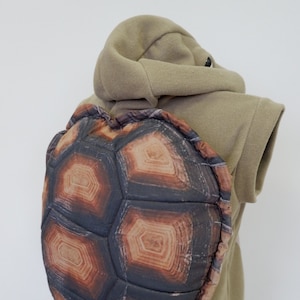 Peut inclure: Un costume de carapace de tortue marron et beige pour enfants. La carapace est en tissu doux et présente un design réaliste. Le costume comprend également un gilet à capuche beige.