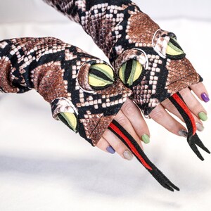 Może przedstawiać: Para brązowo-czarnych fingerlessów z wzorem skóry węża, z czerwonymi i czarnymi akcentami. Rękawiczki mają zielone i żółte oczy oraz czarne rozwidlone języki.