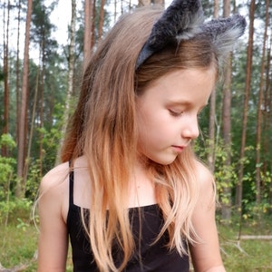 May include: A young girl with long blonde hair wearing a black tank top and a gray furry headband with cat ears. She is standing in a forest.