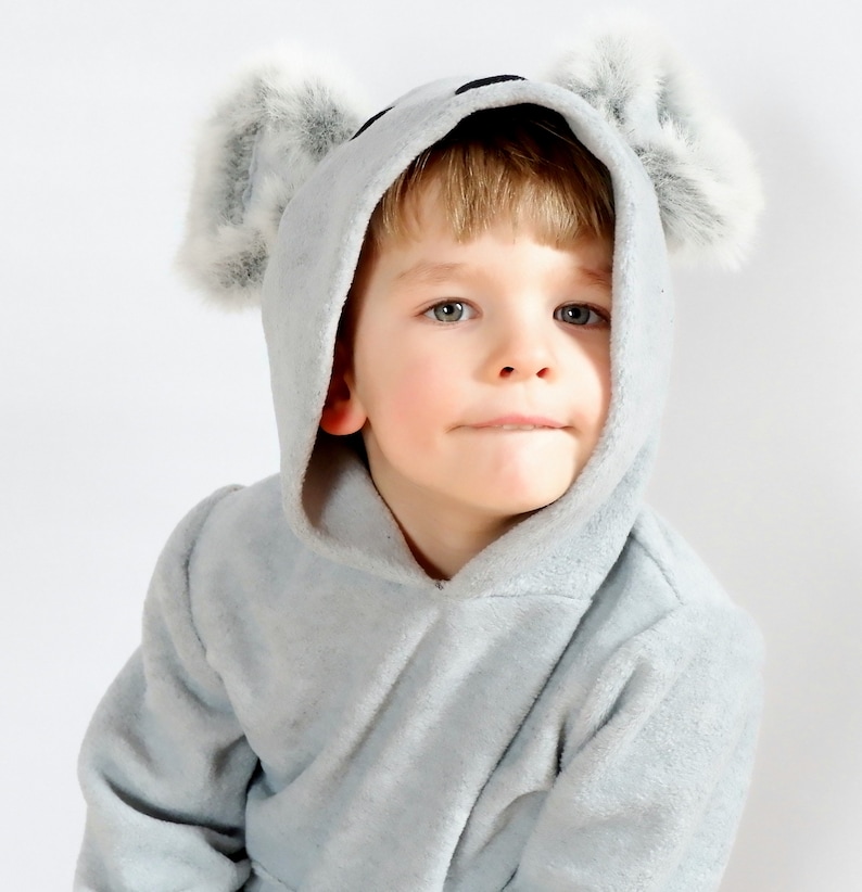 Op de afbeelding: Een jong kind draagt een grijze hooded jumpsuit met fuzzy grijze oren. De jumpsuit heeft een rits aan de voorkant en een capuchon met een trekkoord.