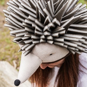 May include: A brown and white hedgehog costume hat with a long snout and black nose. The hat is made of soft, plush fabric and is perfect for a whimsical costume.