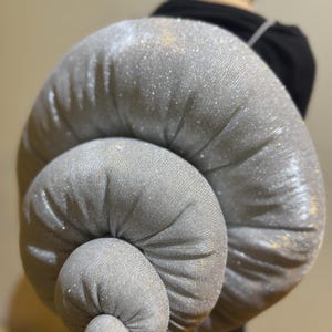 Könnte beinhalten: Ein großes, silbergraues, spiralförmiges Kissen mit einer glitzernden, strukturierten Oberfläche. Das Kissen besteht aus einem weichen Stoff und hat eine geknotete Mitte. Die Gesamtform ähnelt einem Schneckenhaus.