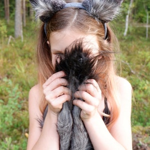 May include: A young person wearing a black dress and a gray wolf ear headband. The headband has soft, fluffy fur. The person is holding the fur of the headband in front of their face.