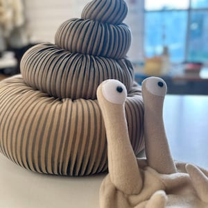 Op de afbeelding: Een pluche slak-speeltje met een grote, spiraalvormige schelp met verticale bruine en beige strepen. Het slakkenlichaam is gemaakt van lichtbeige stof en bevat twee rechtopstaande stelen met witte ronde ogen en zwarte pupillen.